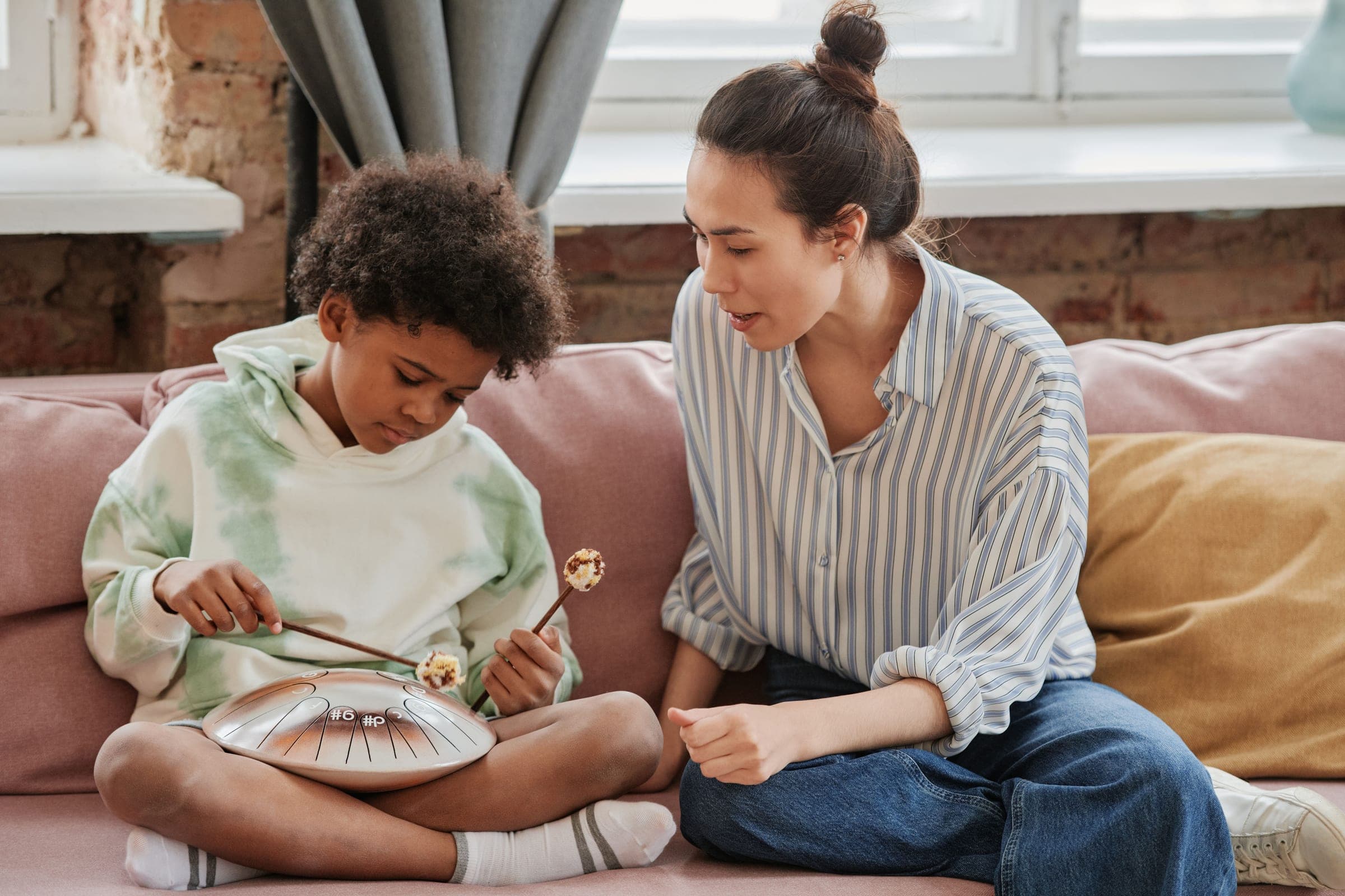 A woman sitting on a sofa beside a young boy, guiding his hands as he plays a small drum