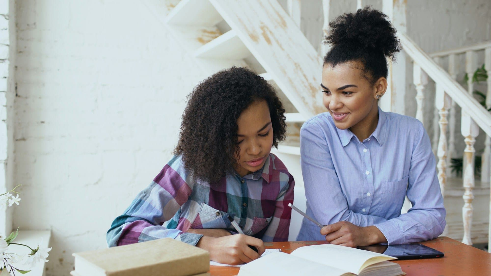 A young woman smiles as she helps a teenage girl work through her homework at a table