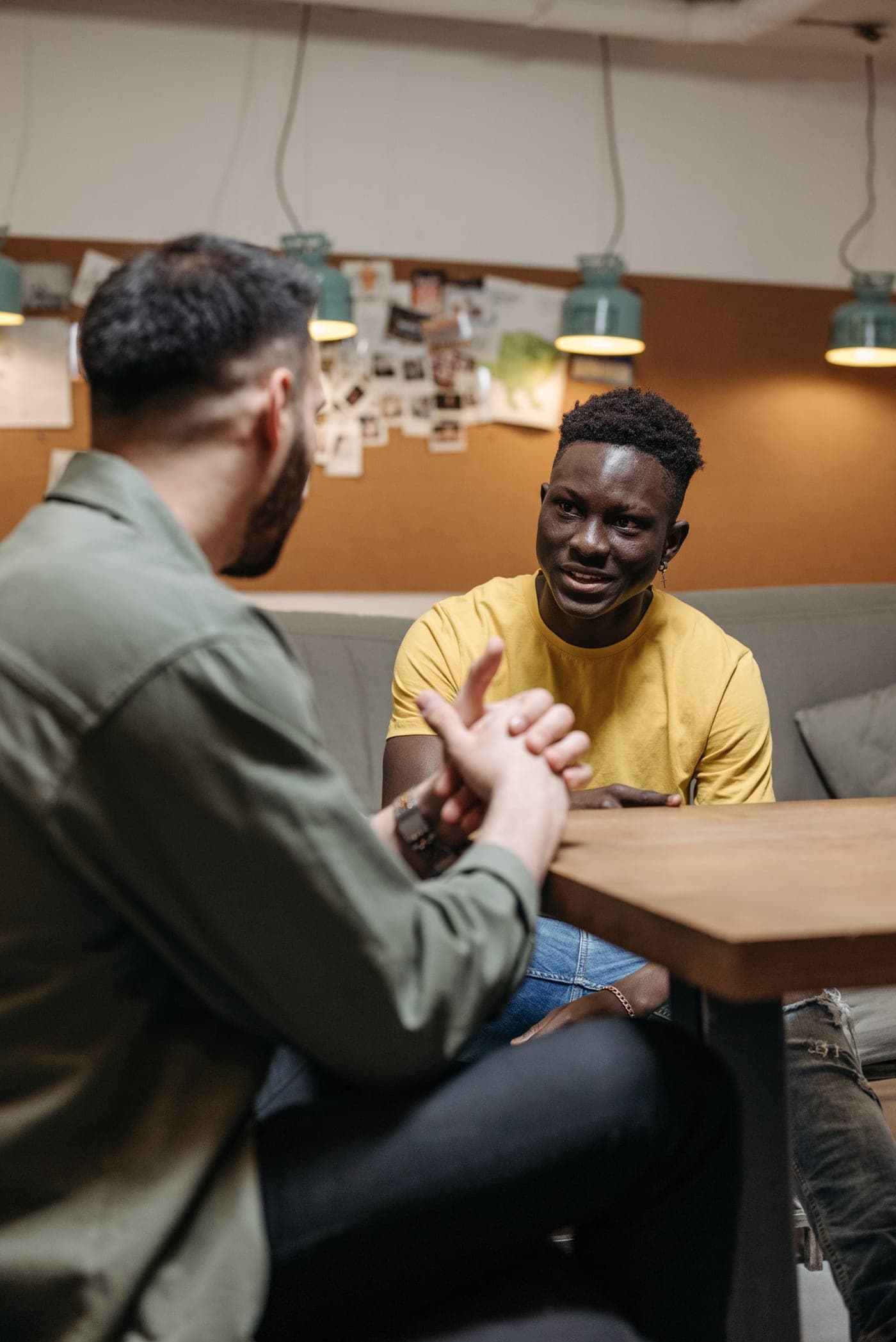 A man leans in to encourage another man as they talk across a table in warm light