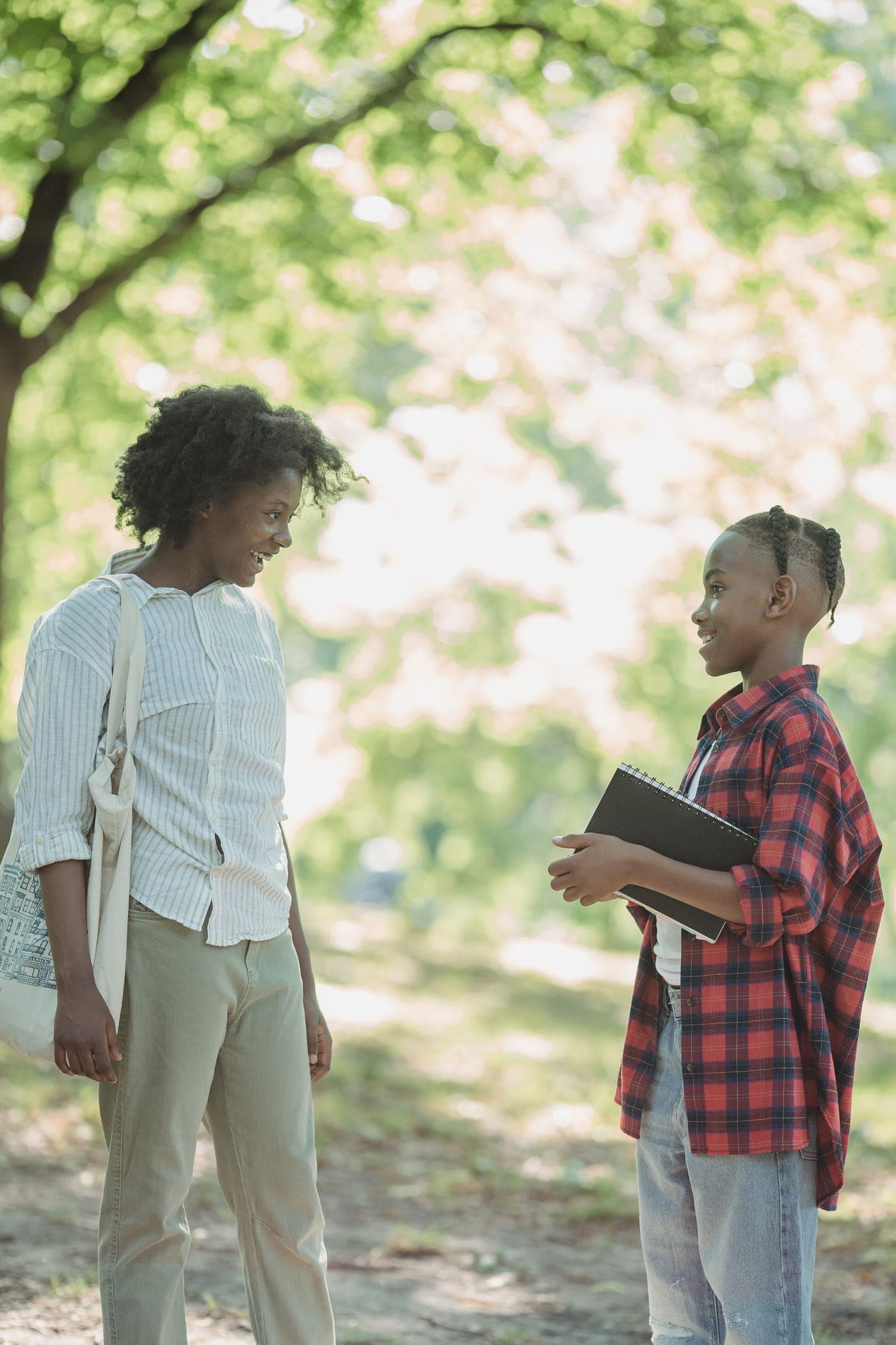 A woman and a boy holding a notebook smile as they talk in a sunny park