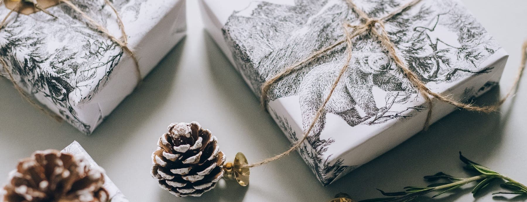 Two gifts wrapped in patterned paper and tied with twine, laid out from above with pine cones and a sprig of greenery