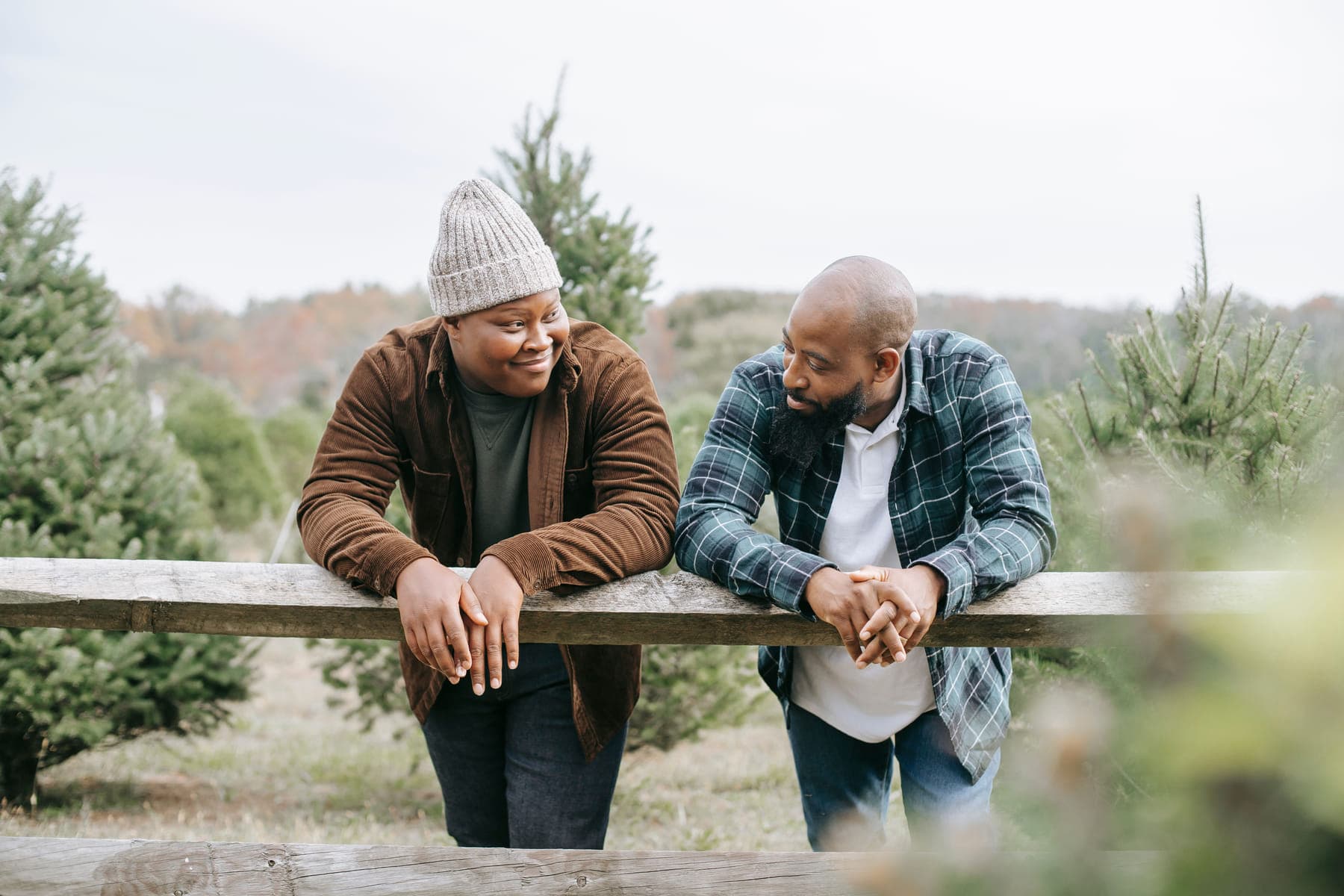 Two people leaning on a wooden fence in an open field, turned toward each other mid-conversation