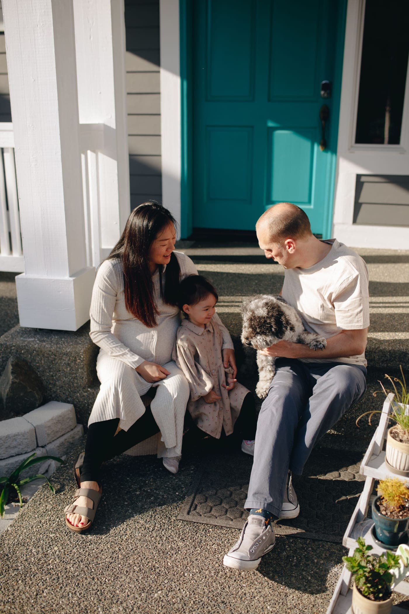 A family sitting together on their front porch steps in the sunshine, the parents either side of a young child holding a small dog