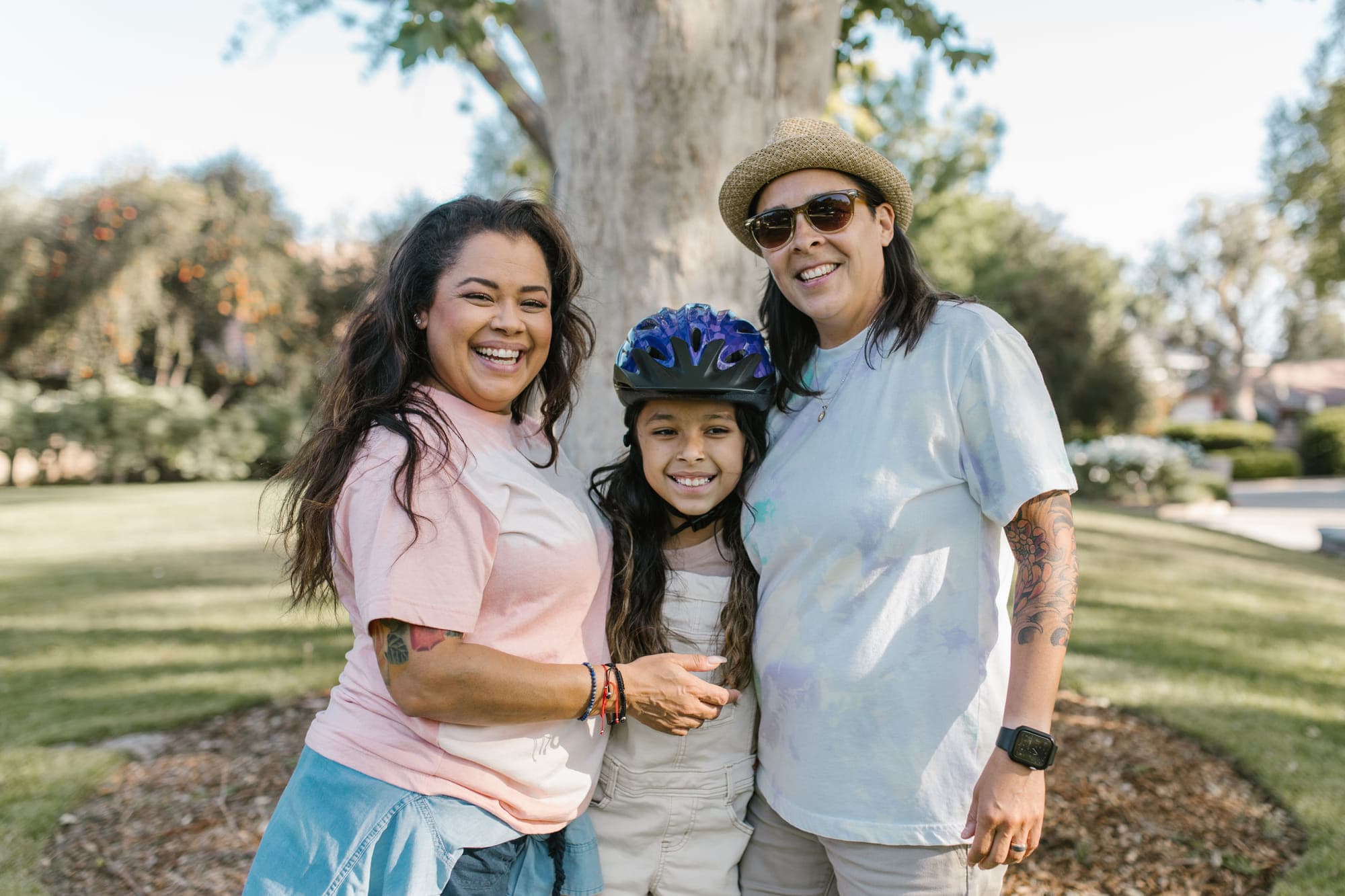Two adults standing either side of a smiling child in a bicycle helmet, outdoors under a tree on a sunny day